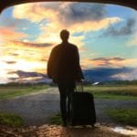 Destinations for Day Trips: Man with a suitcase stands with his back to the camera and looks out at an open field and the setting sun