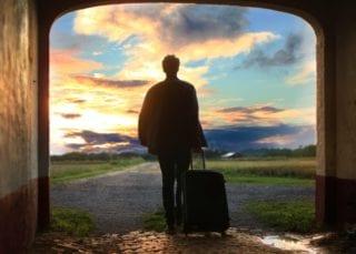 Man with a suitcase stands and looks out at an open field and the setting sun