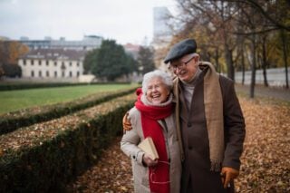 Happy senior couple on walk outdoors in town park in autumn, embracing and laughing