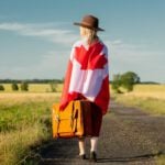Girl in Canada flag with suitcase on country road in sunset