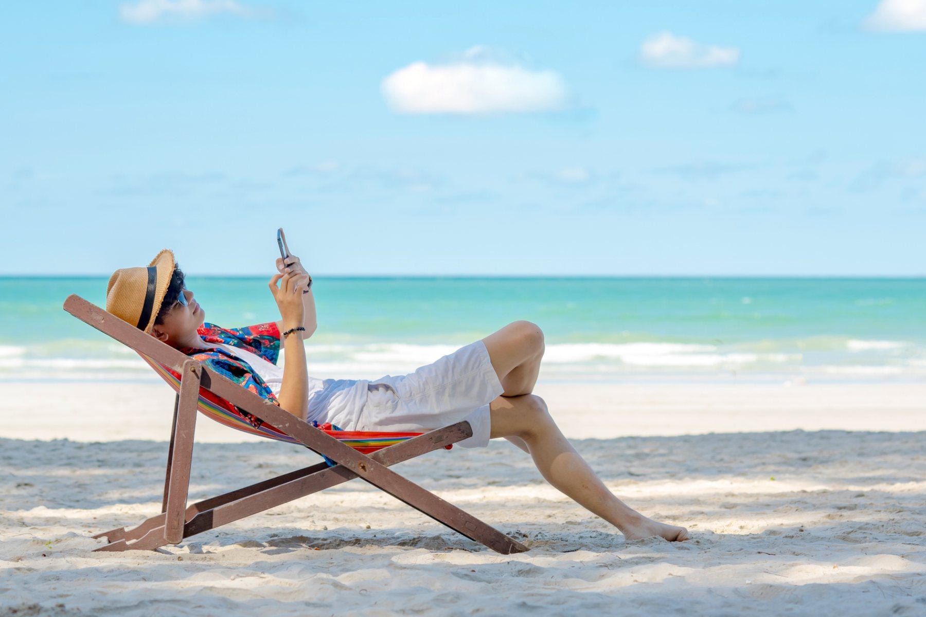 a male expat relaxing in a hammock and using his cell phone on a tropical beach in Thailand, researching the best international credit cards for living abroad