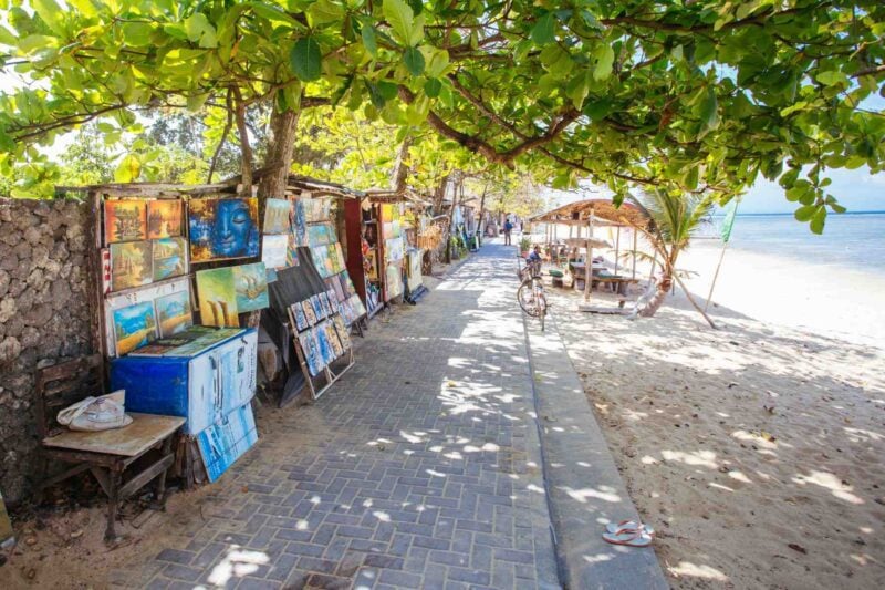 a selection of colorful paintings lined up along a beach in Sanur, Bali