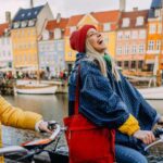Two women laughing and cycling in Denmark, enjoying exercise and sightseeing, promoting a healthy lifestyle while abroad as part of their international healthcare goals